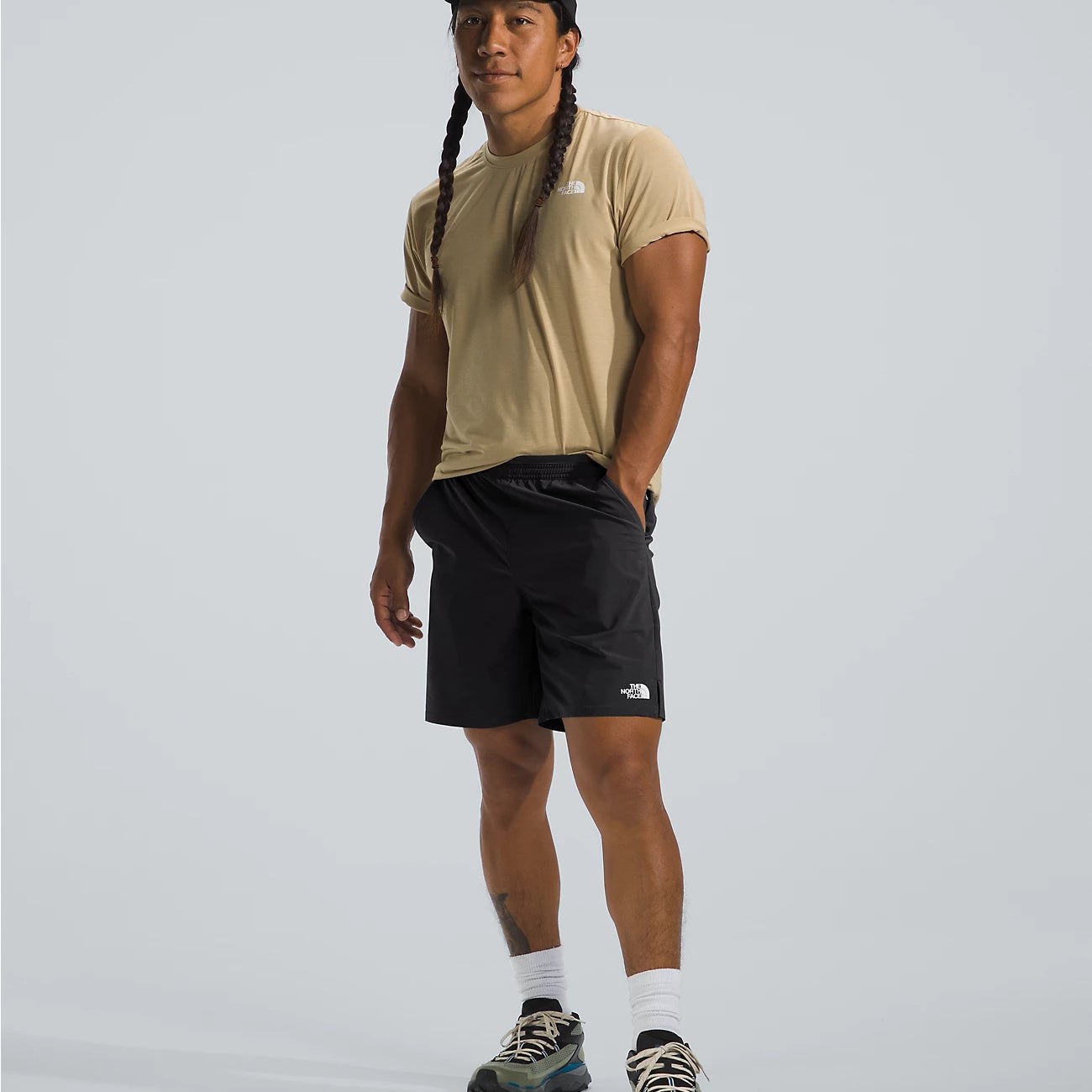 Person wearing a beige t-shirt, black shorts, and a cap on a light gray background