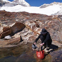 Person collecting water from a mountain stream with snow-capped peaks in the background