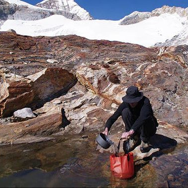 Person collecting water from a mountain stream with snow-capped peaks in the background