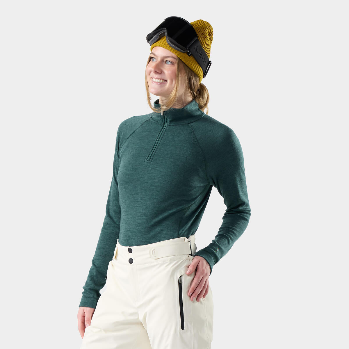 Person wearing a green long-sleeve top and white pants with a yellow beanie and goggles on a light gray background